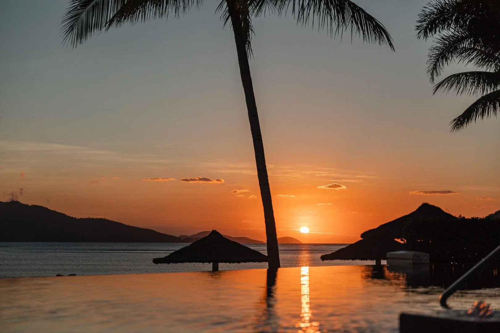 Sunset over the ocean viewed from a tropical beach with palm trees, straw umbrellas, and calm water, with mountains in the background. Travel photographer, Laurene Bardsley Photography, Toowoomba Photographer