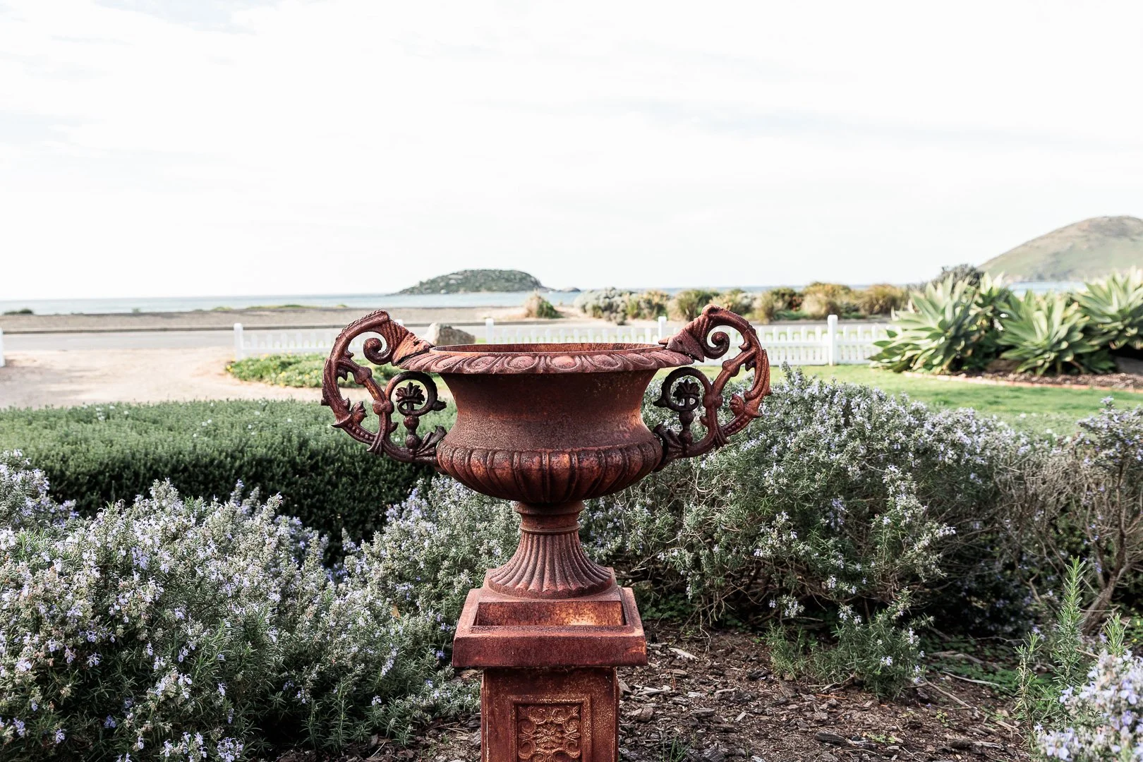 A decorative metal urn on a pedestal in a garden with shrubs, overlooking a beach and ocean, with hills in the background.