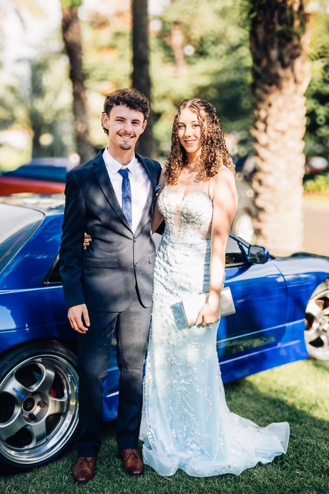 School formal photography couple standing next to a blue sports car outdoors. The man is wearing a dark suit with a blue tie, and his partner is in a blue lace gown holding a clutch. They are smiling with trees in the background.