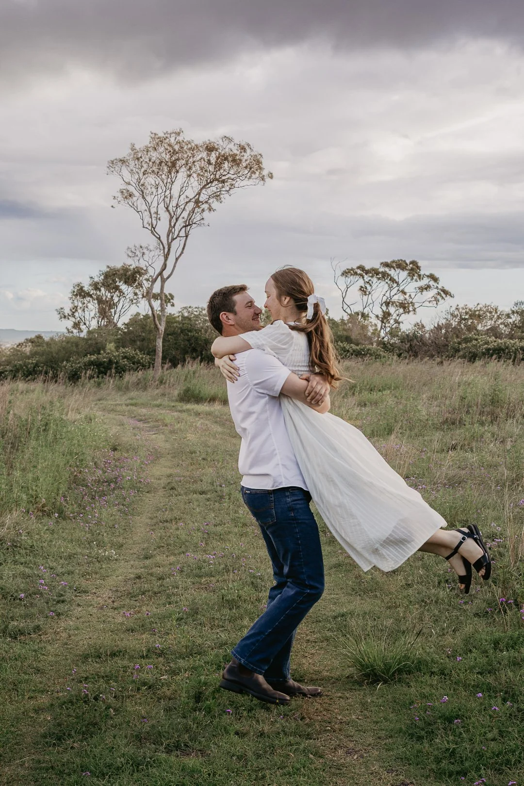 A man lifting a woman in a white dress on a grassy field with trees and overcast sky in the background.