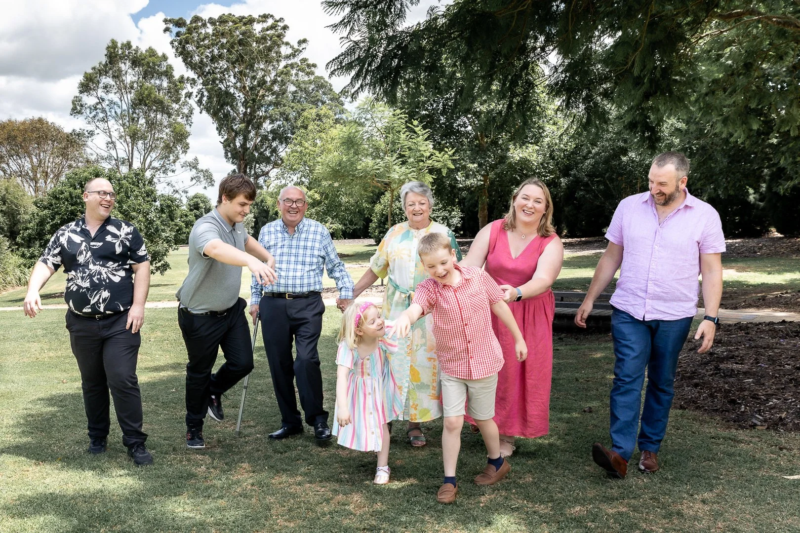 A group of nine people, including children and adults, walking and laughing outdoors in a park with trees and grass.
