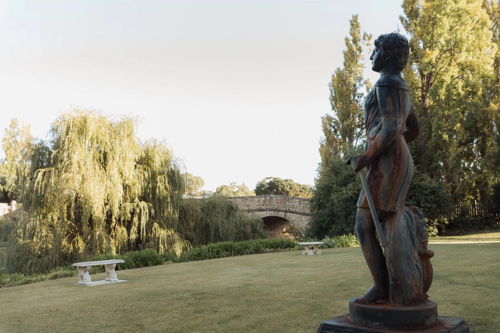 A bronze statue of a young girl holding a stick, standing on a grassy area with trees, a stone bridge, and benches in the background. Design photography, Laurene Bardsley Photography