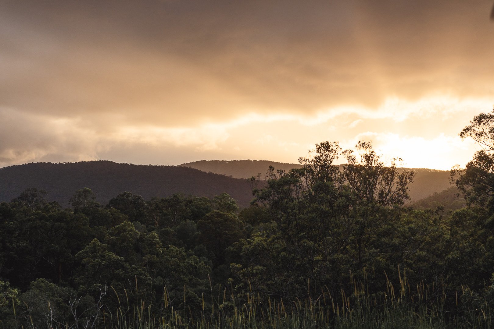 Sunset over a hilly forest with dark green trees and a cloudy sky.