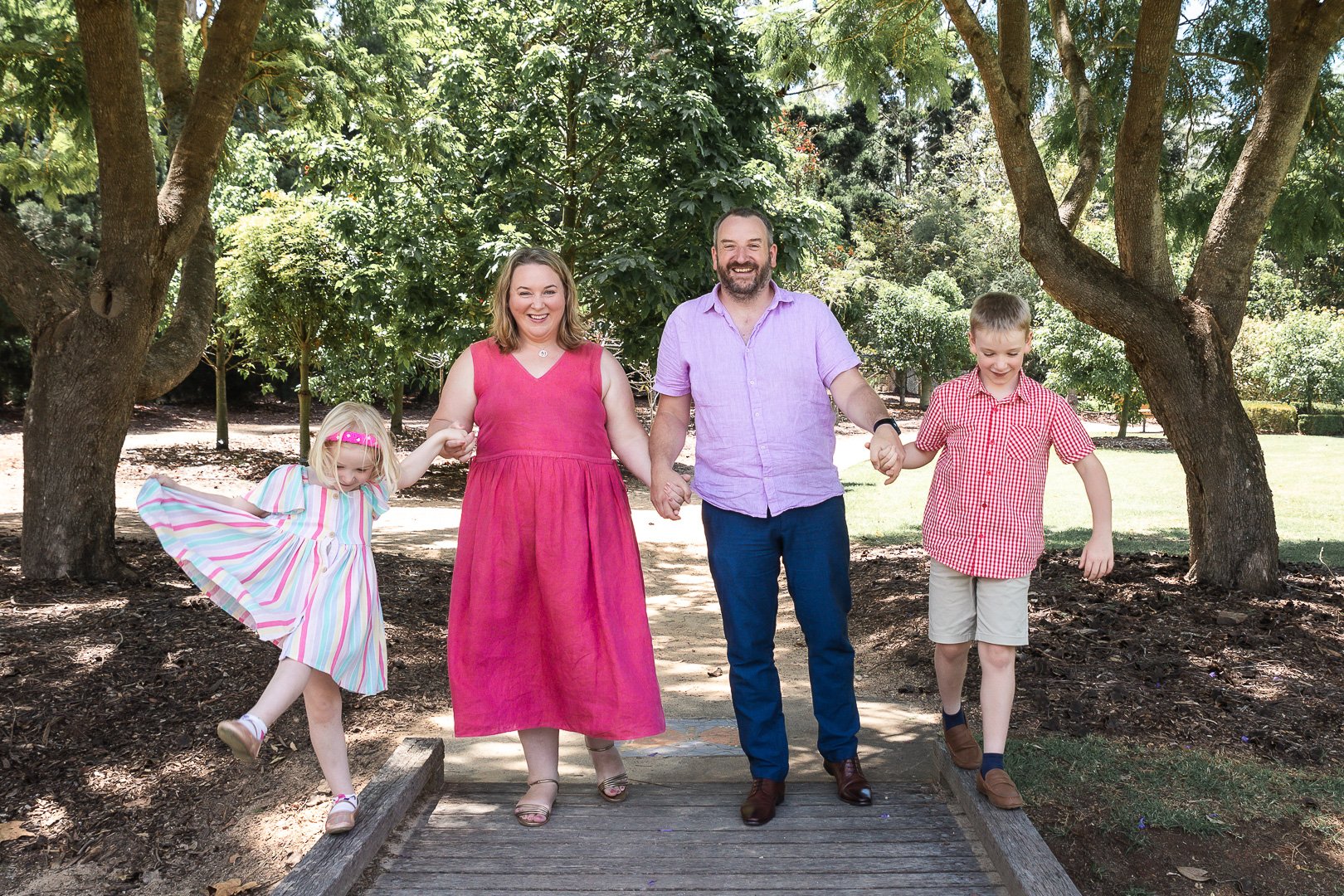 A family of four holding hands and walking on a wooden bridge in a park with lush green trees in the background. The woman is wearing a red dress, the man a purple shirt, the young girl is in a colorful striped dress, and the boy is in a red checkere