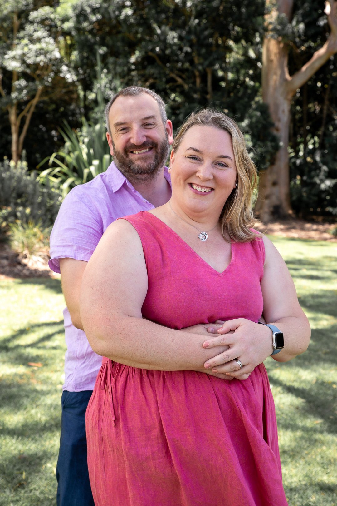 A happy couple standing outdoors in a park or garden, smiling at the camera. The woman is wearing a pink sleeveless dress and a smartwatch, while the man behind her is dressed in a light purple shirt.