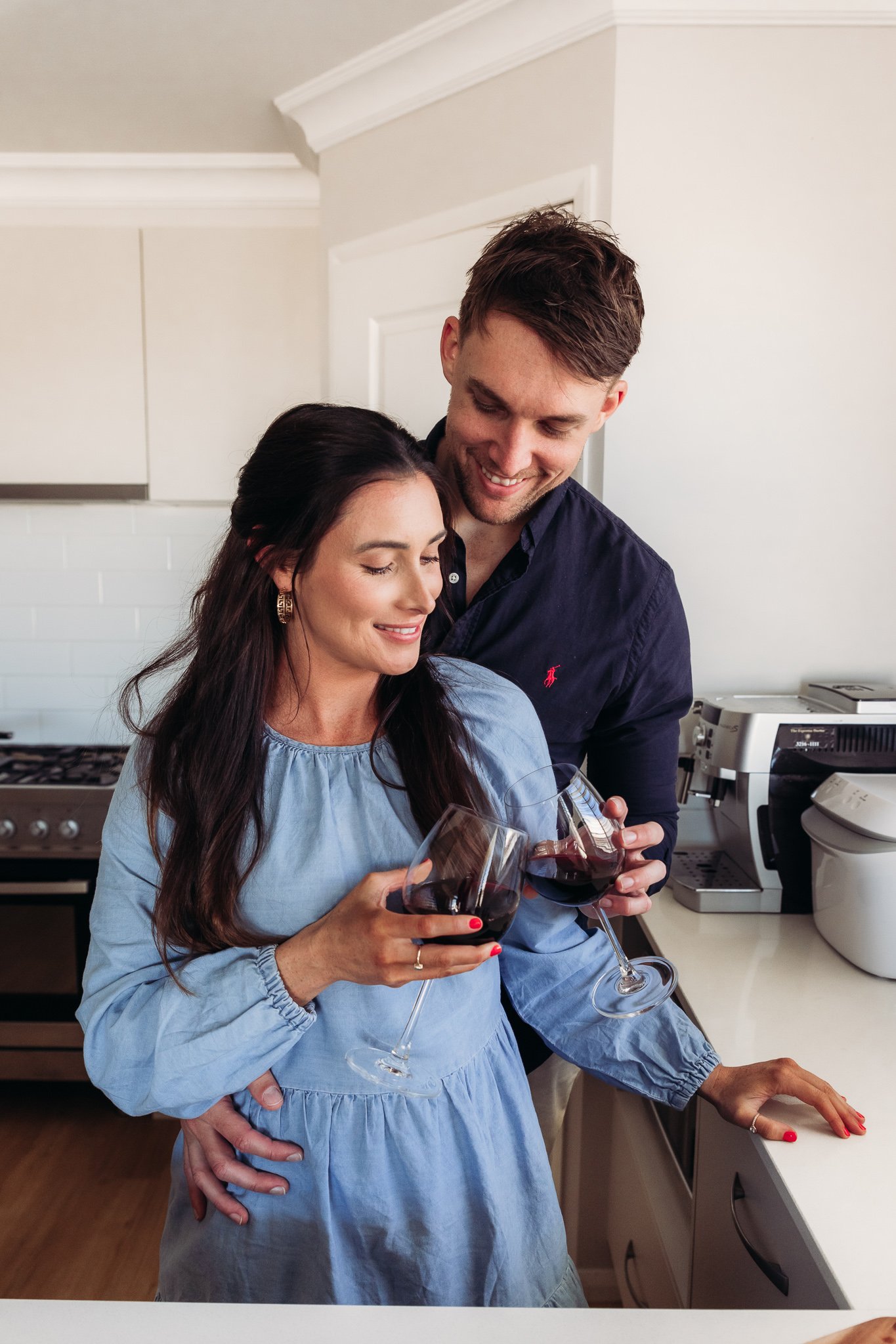 A couple in a kitchen, smiling and holding glasses of red wine.