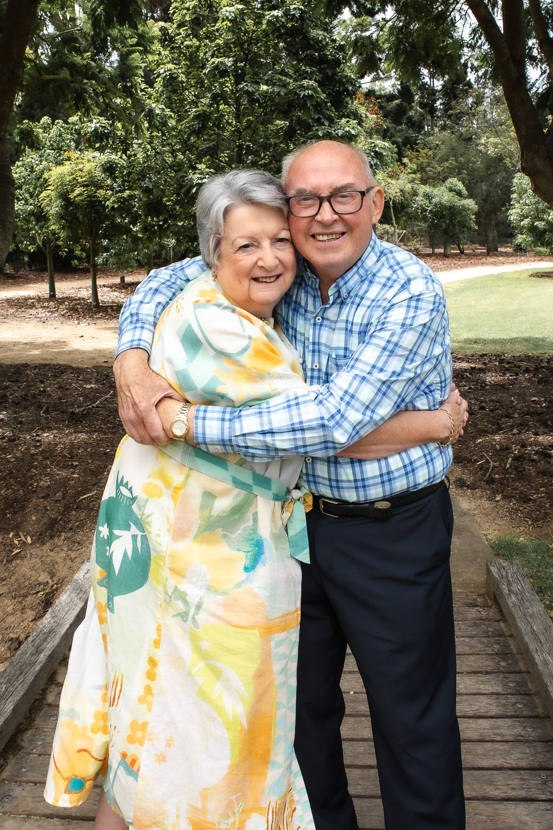 An elderly woman and a middle-aged man are hugging and smiling on a wooden bridge in a park with green trees in the background.