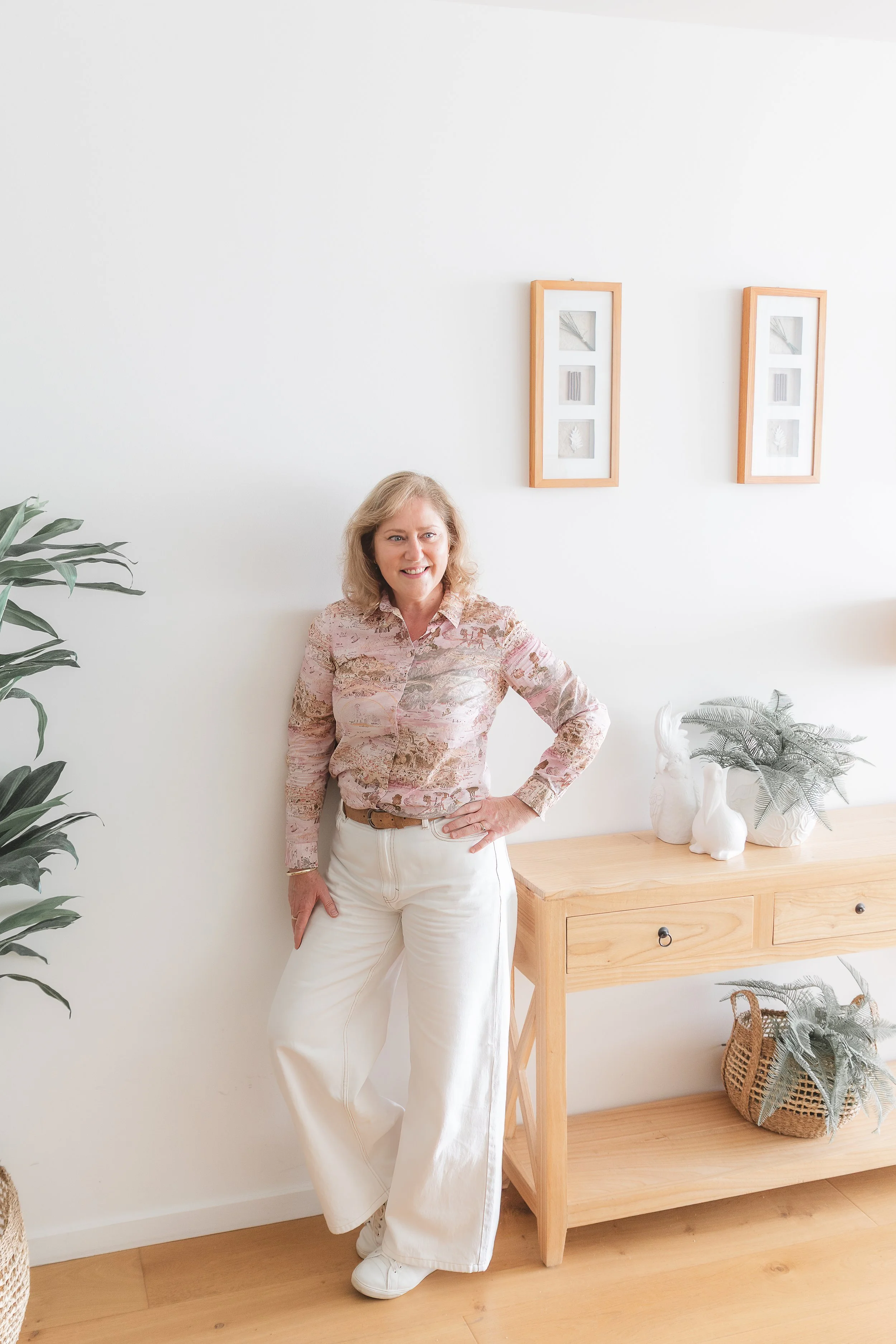 A woman with blonde hair wearing a pink patterned blouse, white pants, and white sneakers, standing in a bright, minimalistic room near a wooden side table with white ceramic animal and plant decor, and framed botanical art on the white wall.