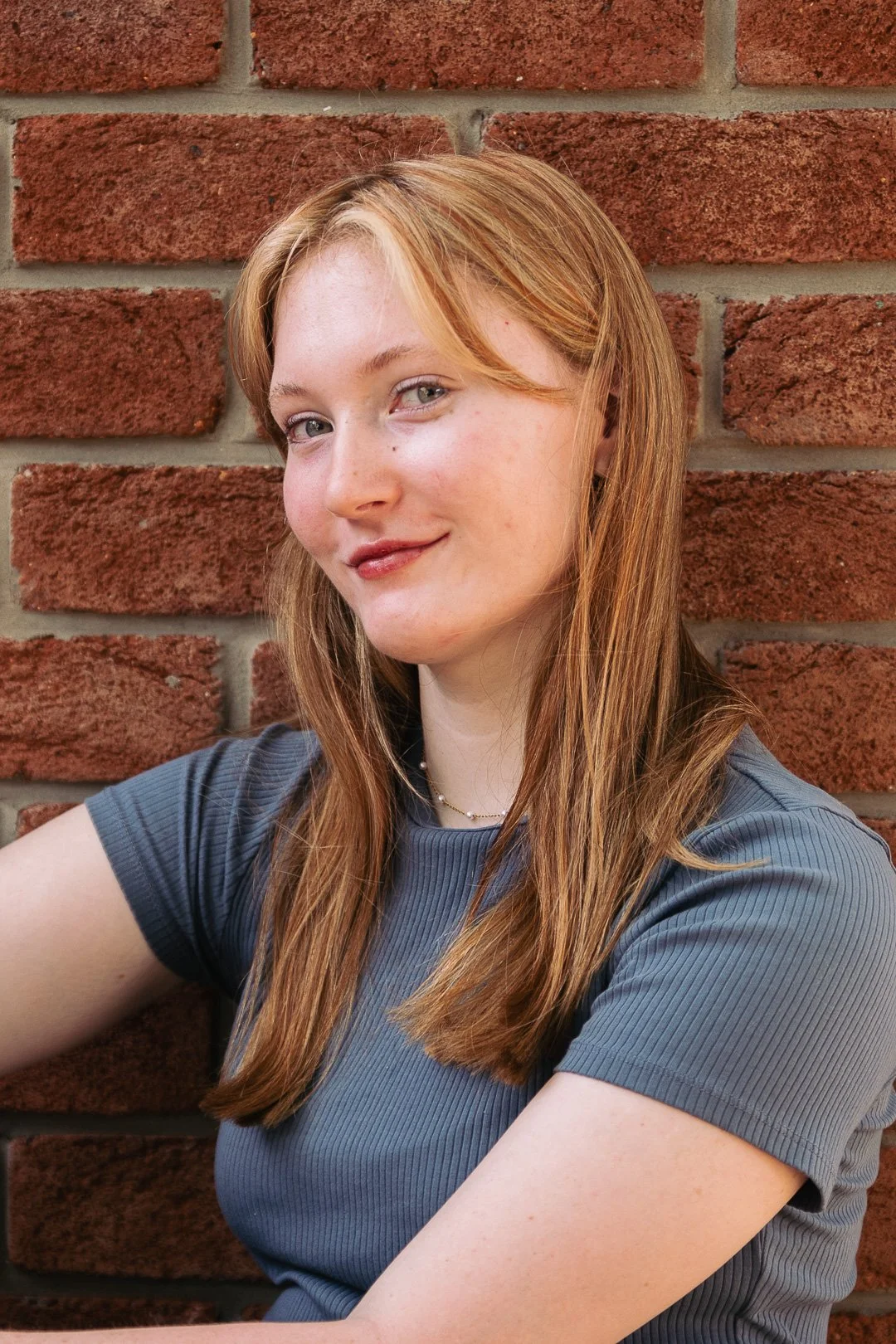 A young woman with red hair and blue eyes is smiling slightly while leaning against a red brick wall, wearing a gray shirt.