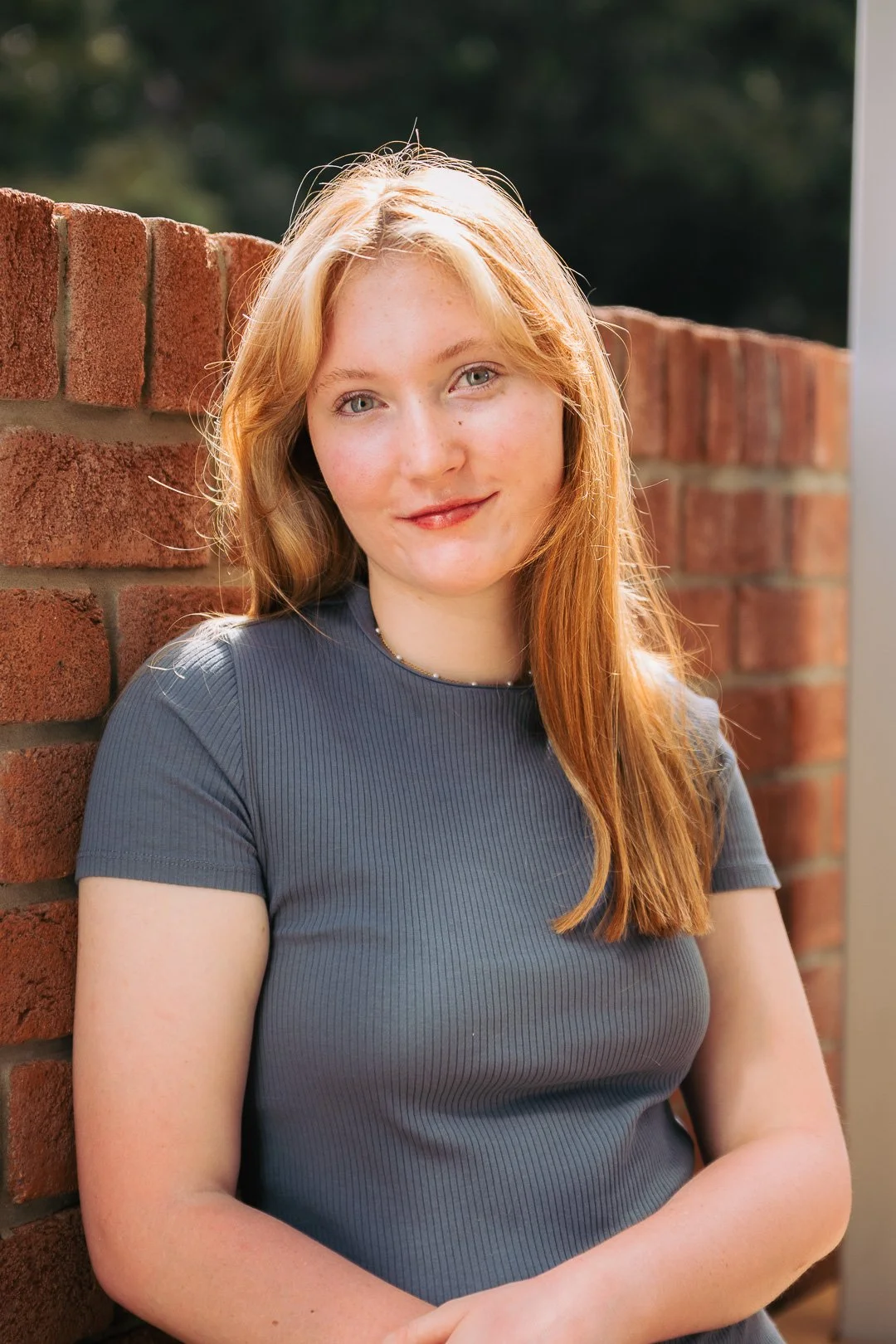 A young woman with red hair, wearing a gray top, leaning against a brick wall outdoors on a sunny day.