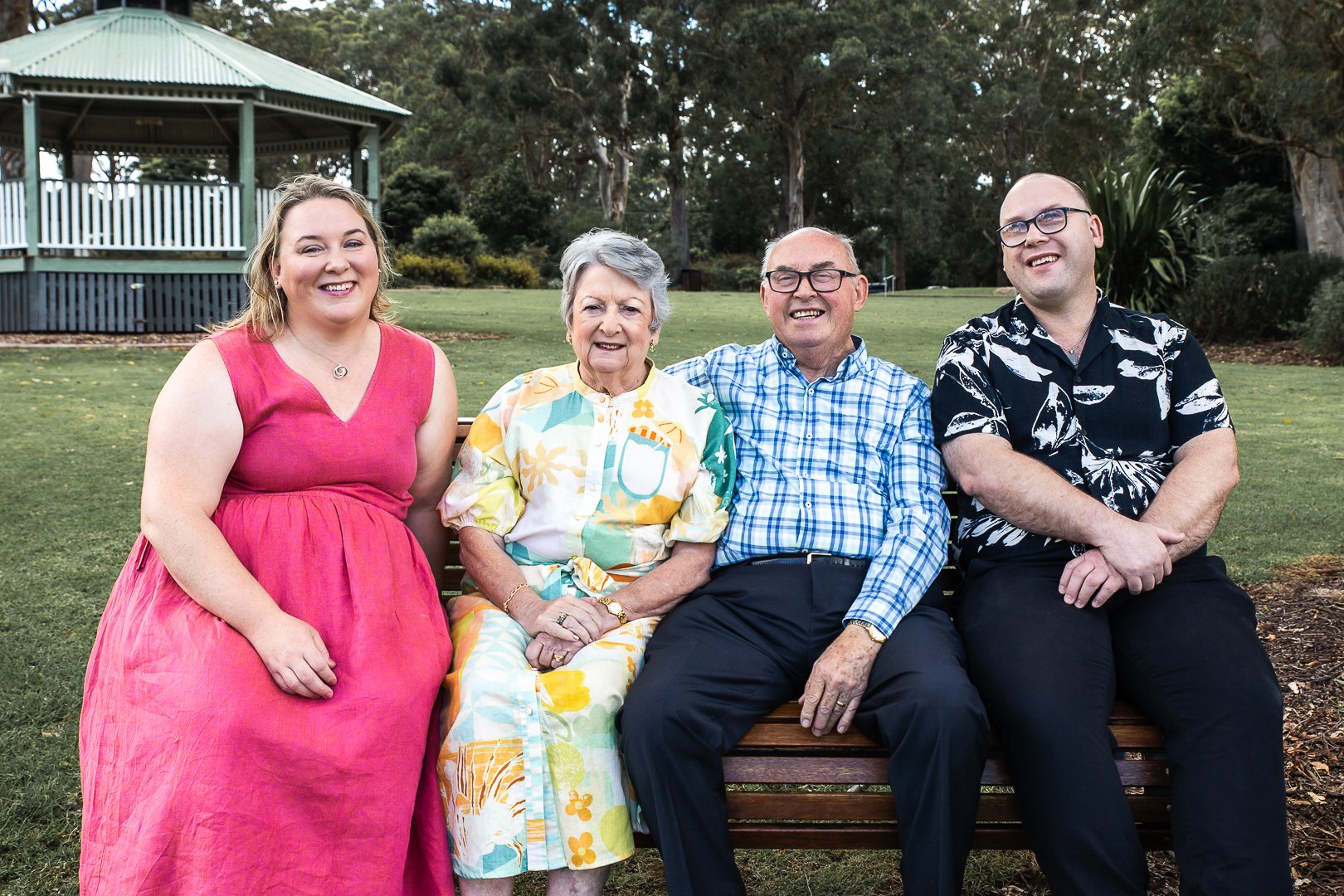 Four people sitting on a park bench, smiling, with trees, grass, and a pavilion in the background.