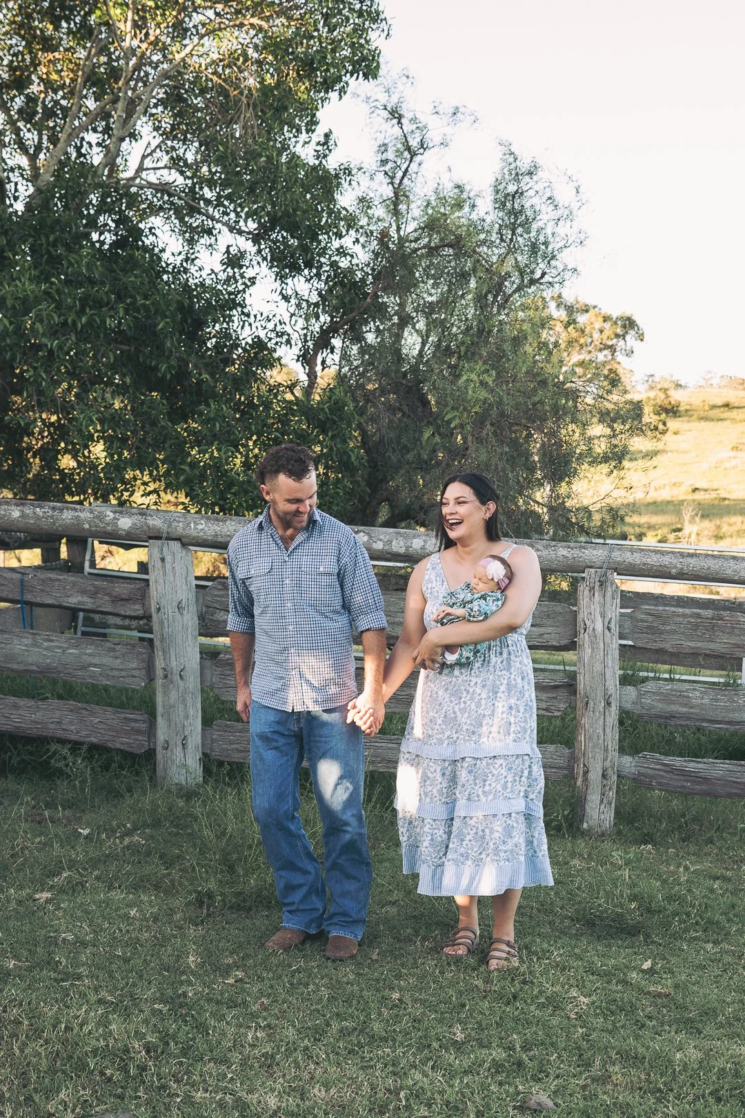 A family of three standing outdoors in front of a wooden fence, with trees and a grassy field in the background. The woman is holding a baby, and all three are smiling and enjoying each other's company.