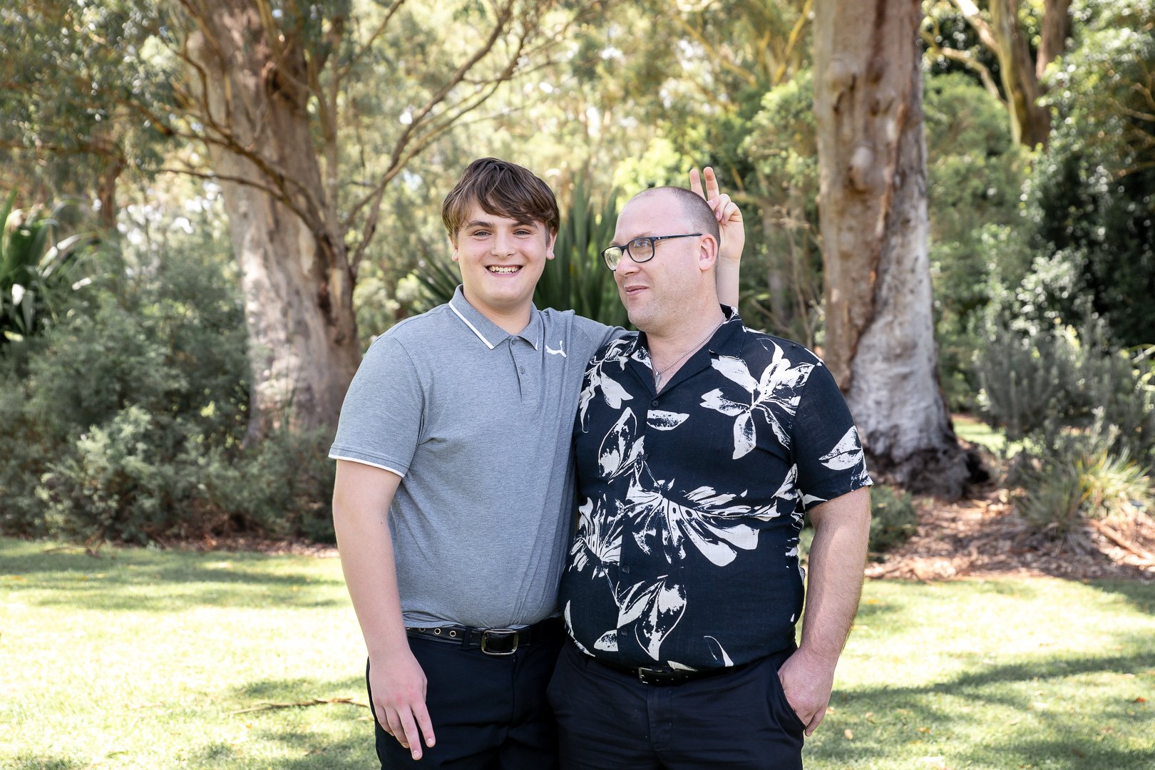 Two men standing outdoors in a park, one appears younger with a gray polo shirt, and the other older with a patterned black and white shirt. The older man is making bunny ears behind the younger man's head.