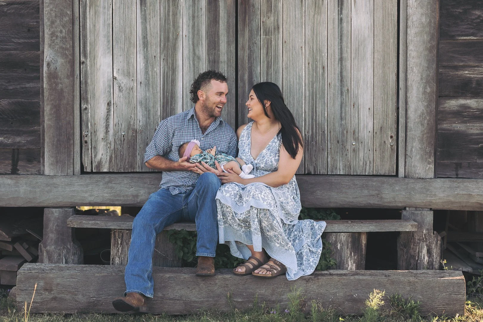 A family of three sitting outdoors on a wooden porch. The father and mother are smiling at each other while holding a newborn baby girl dressed in floral clothing and a purple hat. Family lifestyle photography session.