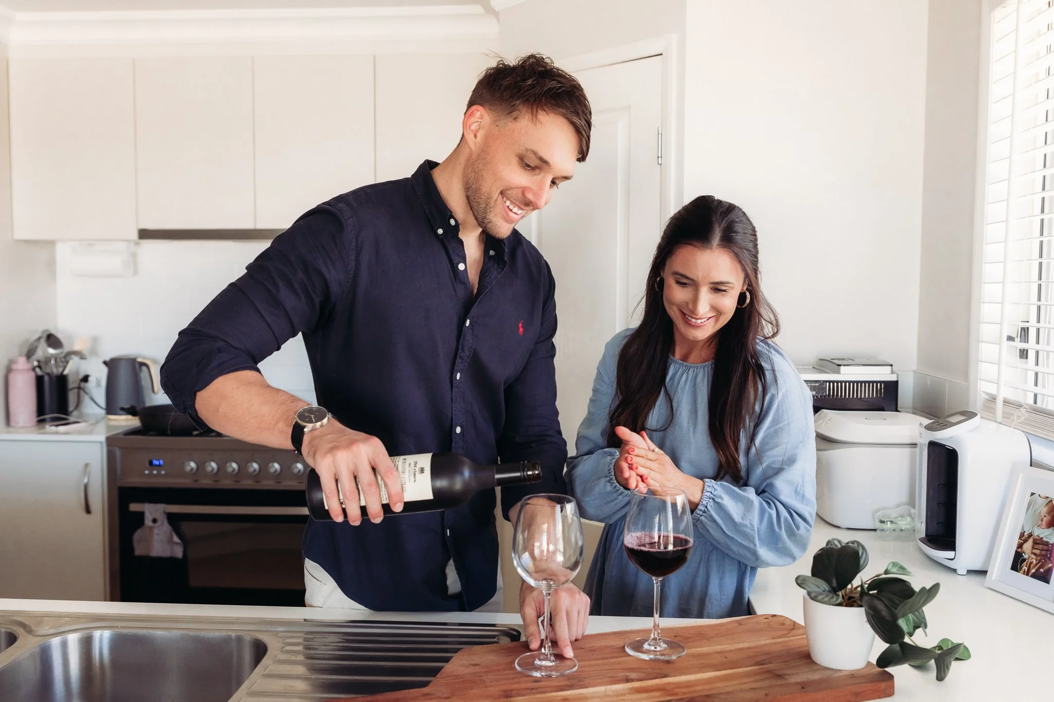 A man pouring red wine into a wine glass while a woman watches and smiles in a bright kitchen.