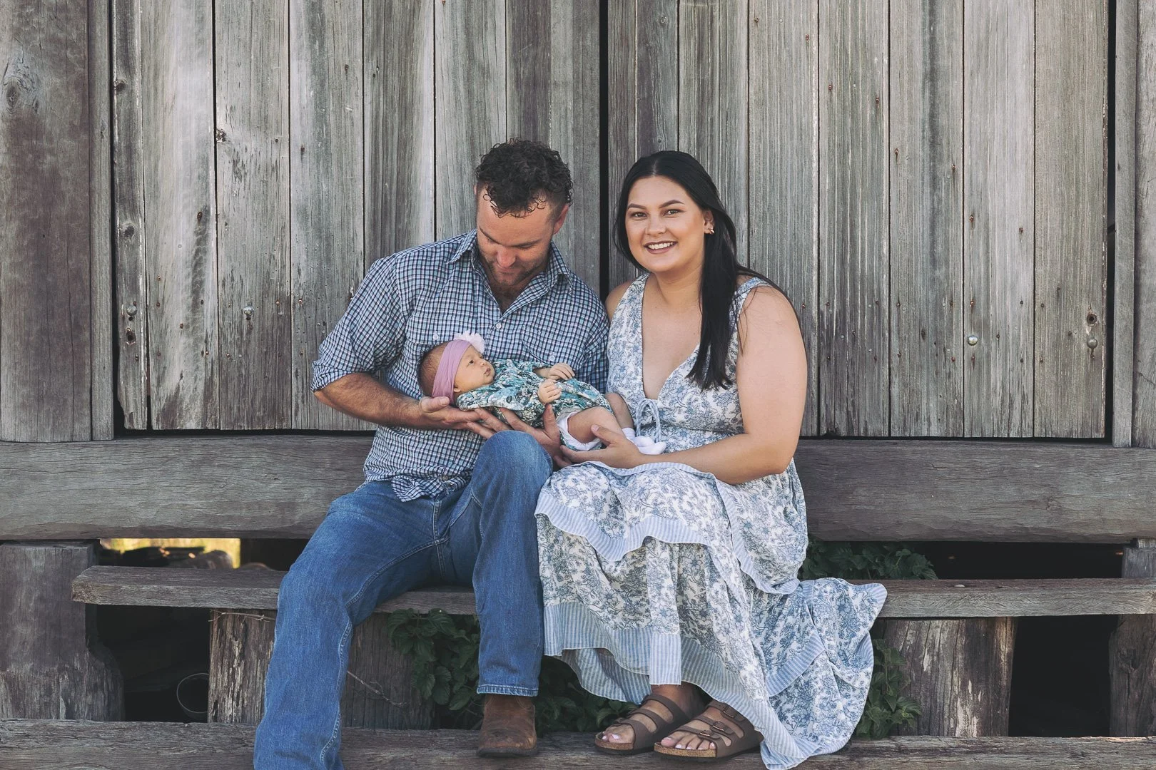 A family of three sitting outdoors in front of a wooden fence. The father and mother are smiling, and the father is holding a newborn baby girl with a pink headband.