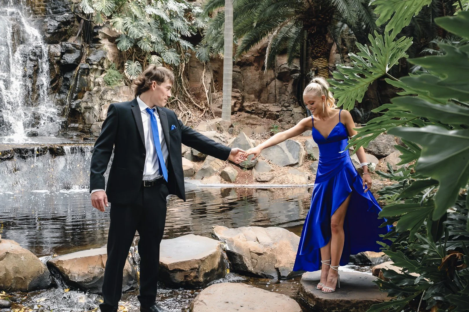 A young man in a black suit and blue tie holds hands with a young woman in a royal blue dress with a high slit, standing on rocks beside a small waterfall and lush greenery during their formal photography session.
