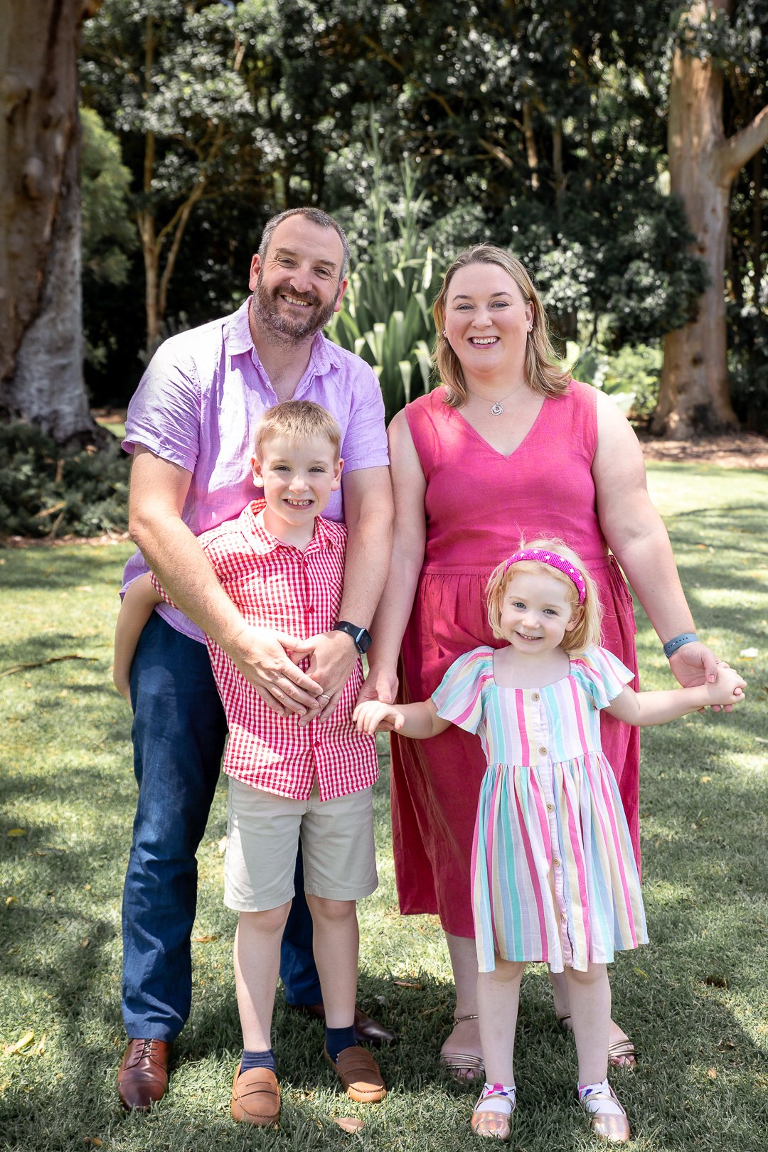 A family of four, including a man, woman, young boy, and young girl, standing outdoors on grass with trees in the background, smiling and holding hands.