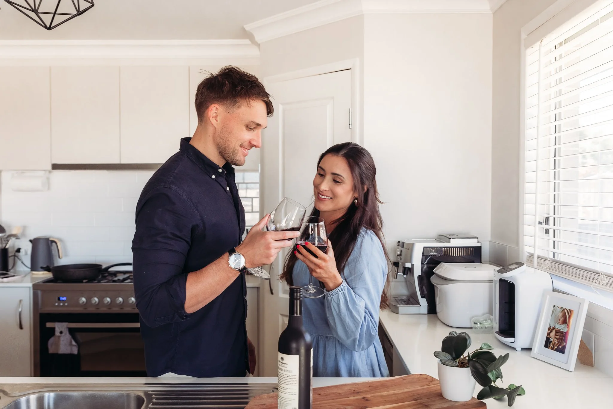 A couple clinking glasses of red wine in a modern kitchen.