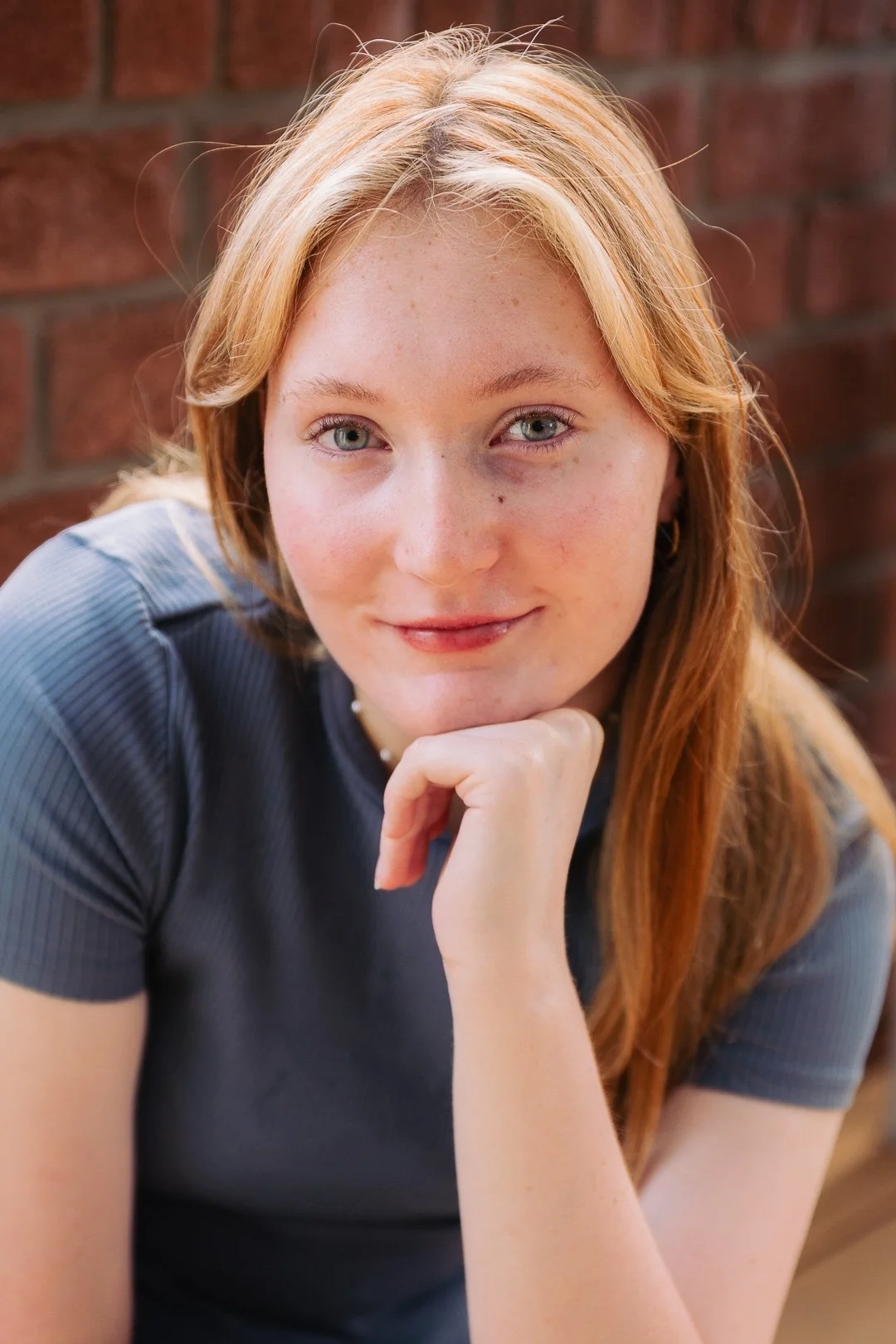 A woman with red hair and light skin, resting her chin on her hand, wearing a gray top, in front of a brick wall.