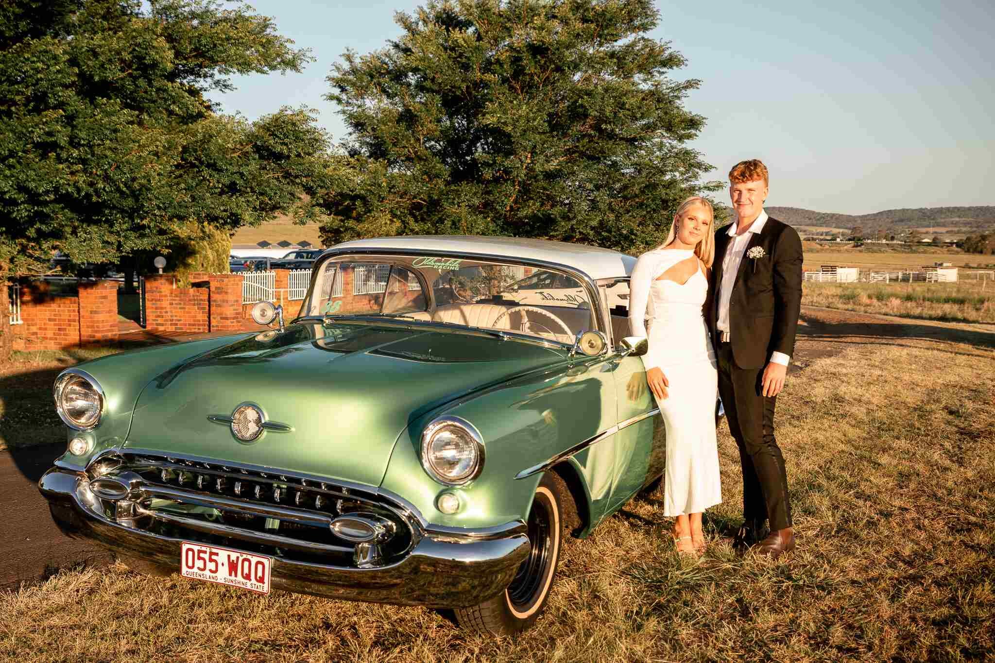 A young couple are celebrating their school formal and posing in front of a green shiny classic car in the late afternoon sunshine.