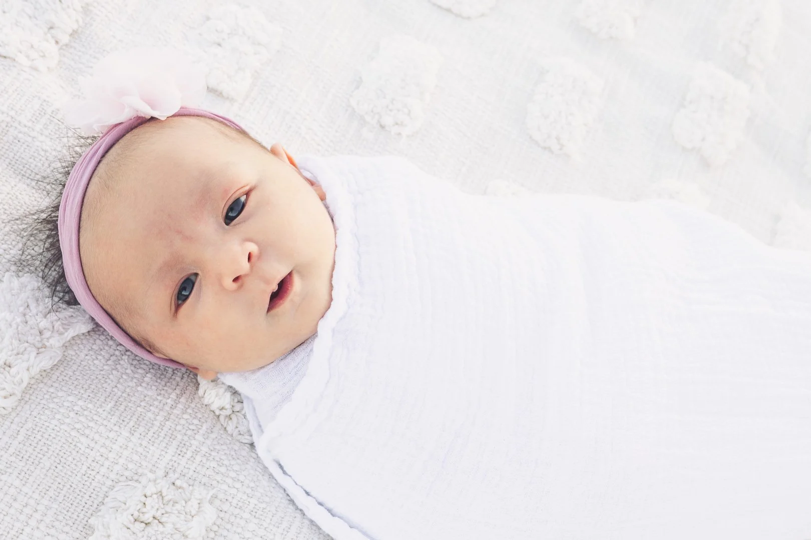 A baby lying on a textured white blanket, wearing a pink headband with a bow, wrapped in a white cloth.