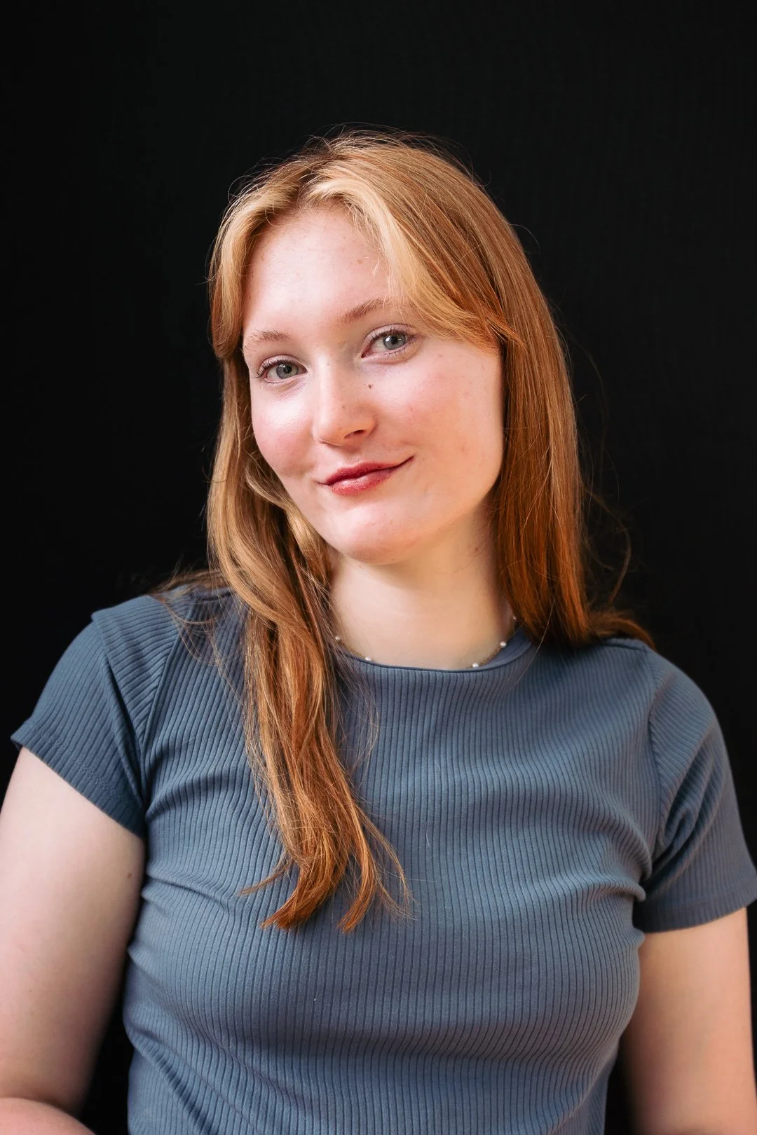 A young woman with red hair smiling at the camera, wearing a blue shirt against a black background.