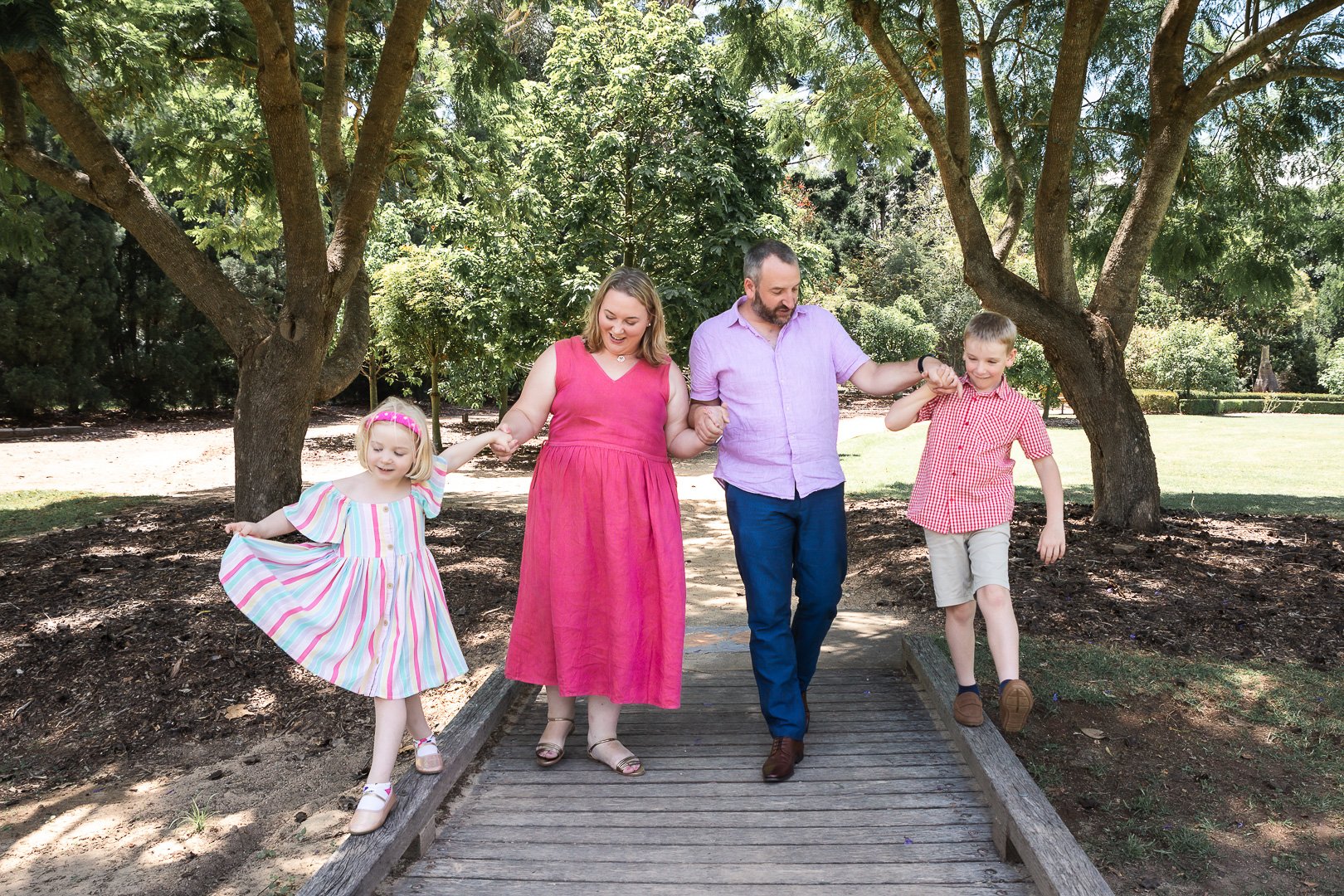 A family of four walking hand-in-hand on a wooden bridge surrounded by trees and greenery, smiling and enjoying a sunny day outdoors.