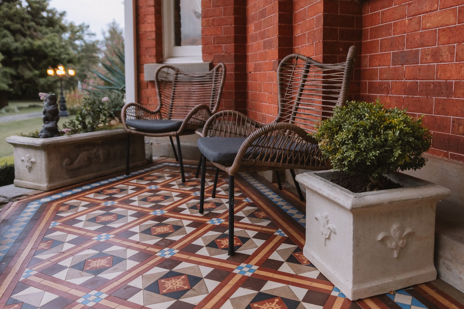 Two outdoor wicker chairs with grey cushions on a mosaic-tiled porch, with a large potted shrub and a brick house wall in the background. Boutique accommodation photographer, Toowoomba. Laurene Bardsley Photography
