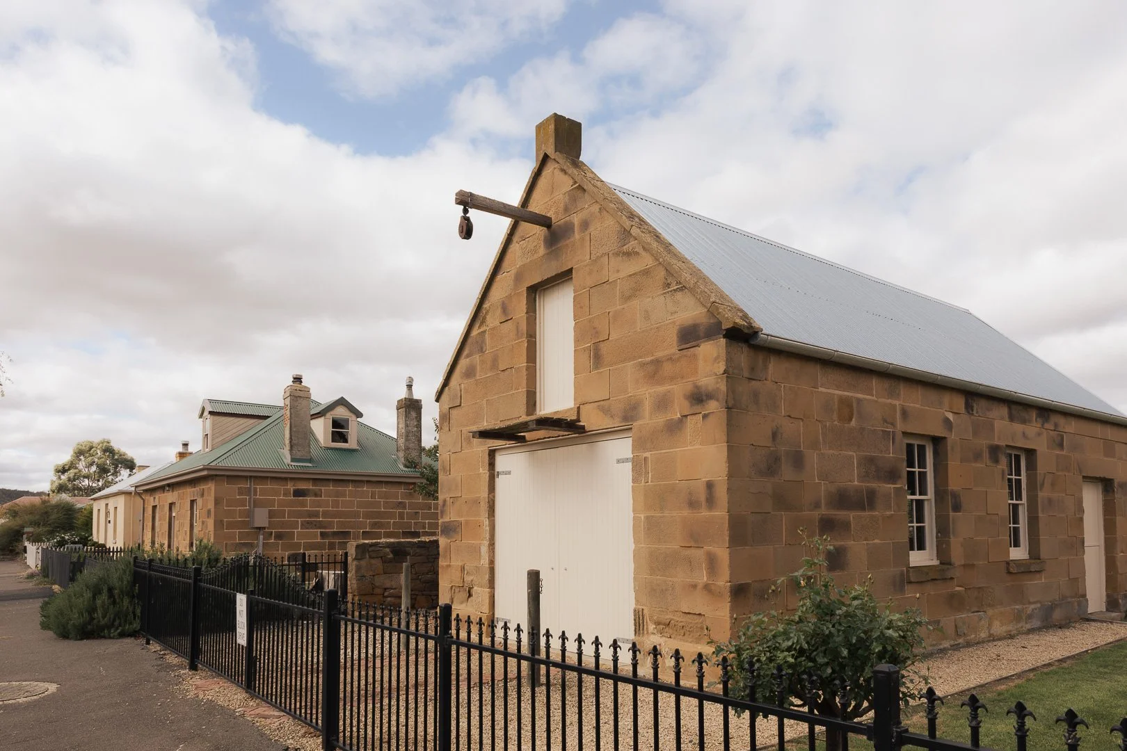 A stone house under construction with a metal roof and a garage door, surrounded by a black iron fence and neighbouring stone houses with green metal roofs. Historic home architectural photography, Toowoomba. Laurene Bardsley Photography.