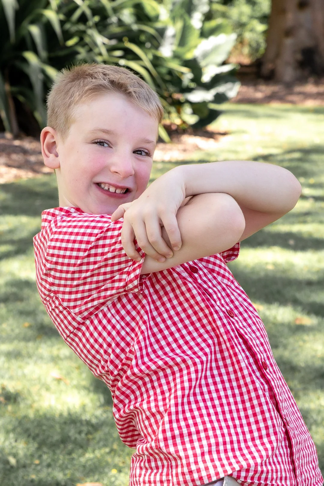 A smiling boy with short blond hair, wearing a red and white checkered shirt, is outdoors in a garden or park, pulling up his sleeve to show his arm.