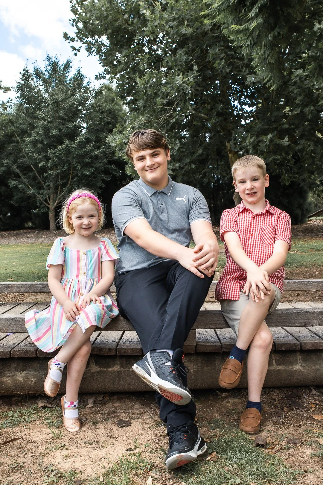 Three children sitting on a wooden park bench outdoors, smiling at the camera with trees in the background.