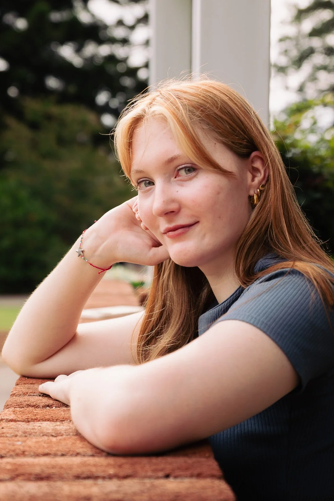 A young woman with shoulder-length red hair and light skin, smiling softly, resting her head on her hand while sitting at a brick wall outdoors with greenery in the background.