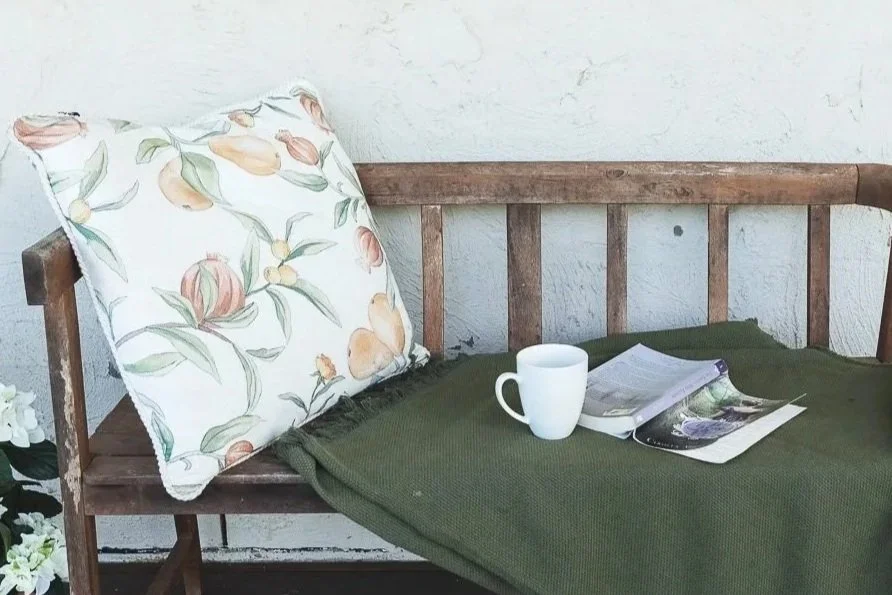 Wooden bench with a floral pillow, a white mug, and open magazines or books on a green blanket, against a textured white wall.