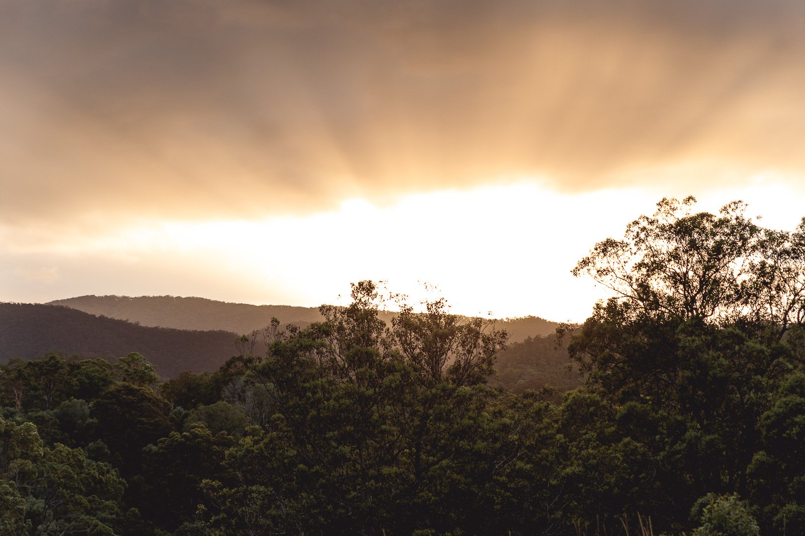 A landscape view of forested hills with a dark storm cloud covering the sky and a bright white light near the horizon.
