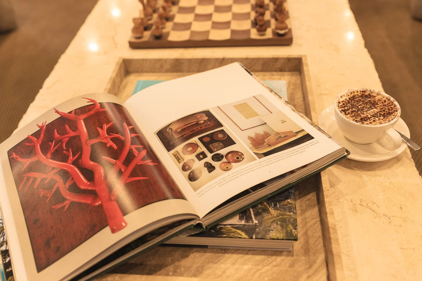 An open art book on a table displaying images of red coral and various art objects. Next to the book is a cup of coffee with chocolate sprinkles on top.