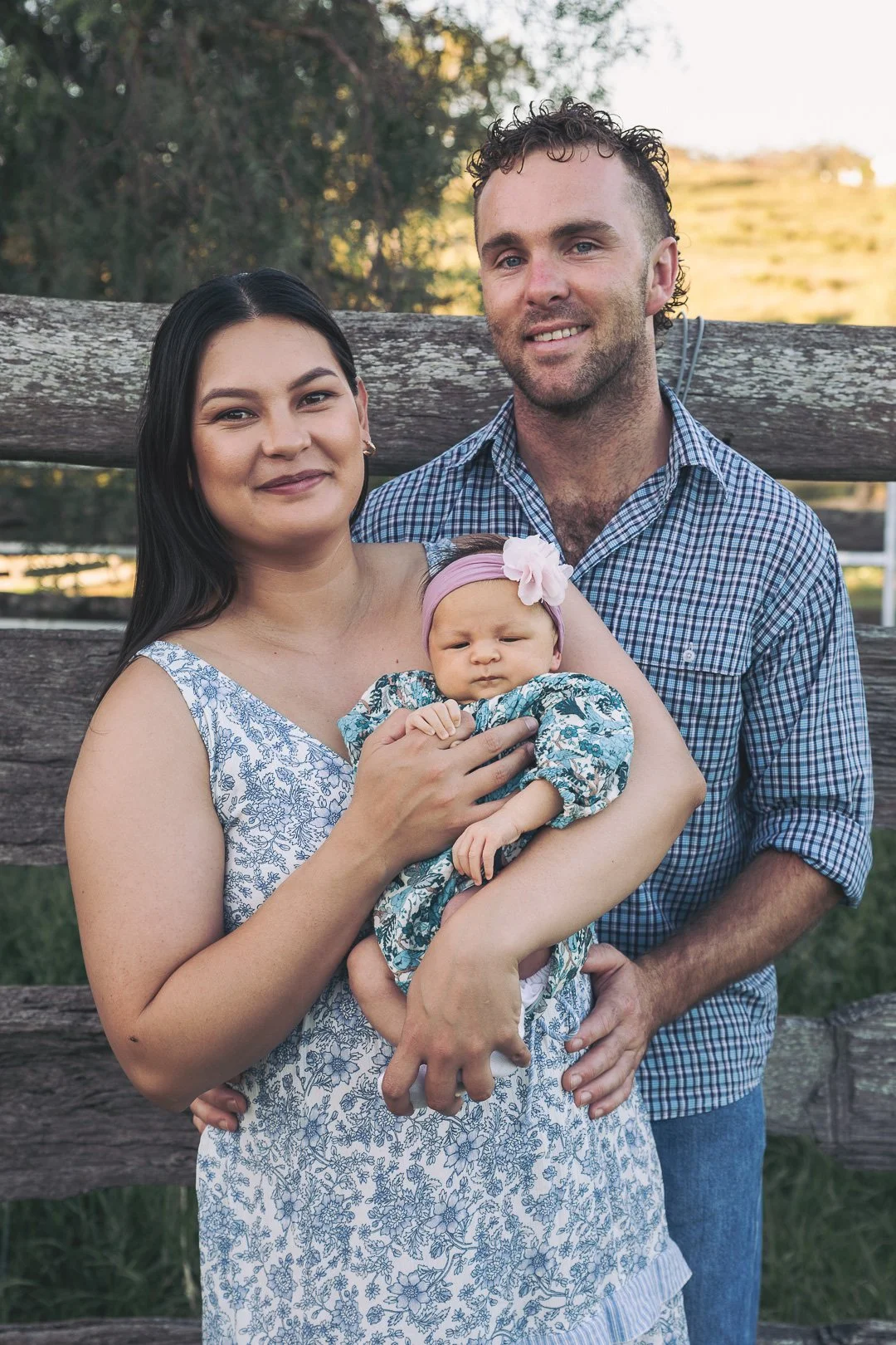 A family of three, two adults and a baby, standing outdoors in front of a wooden fence with trees and hills in the background, during daylight.