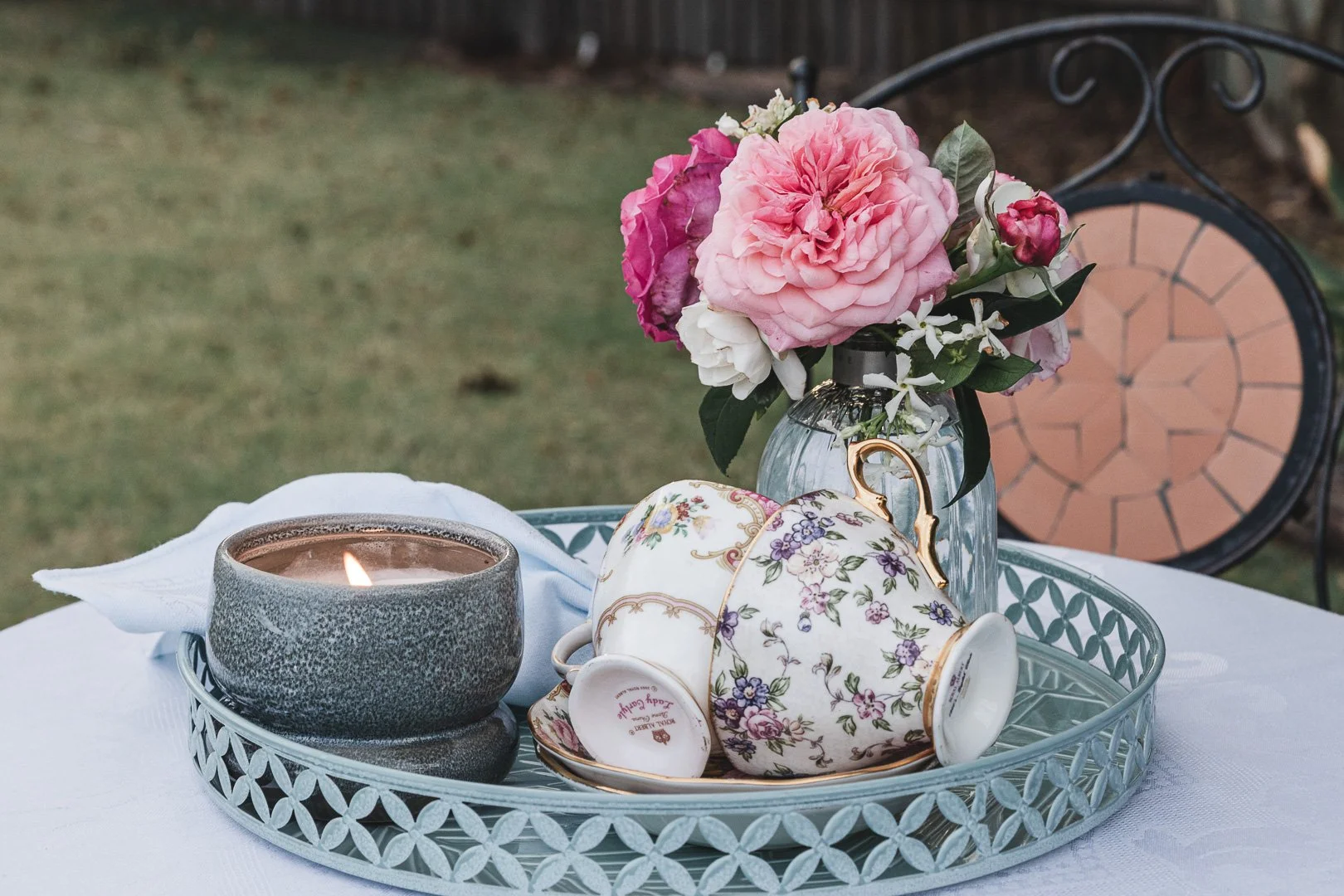 A tray with a lit candle, a floral teacup, and a vase of pink and white flowers on a white tablecloth on a table outdoors.