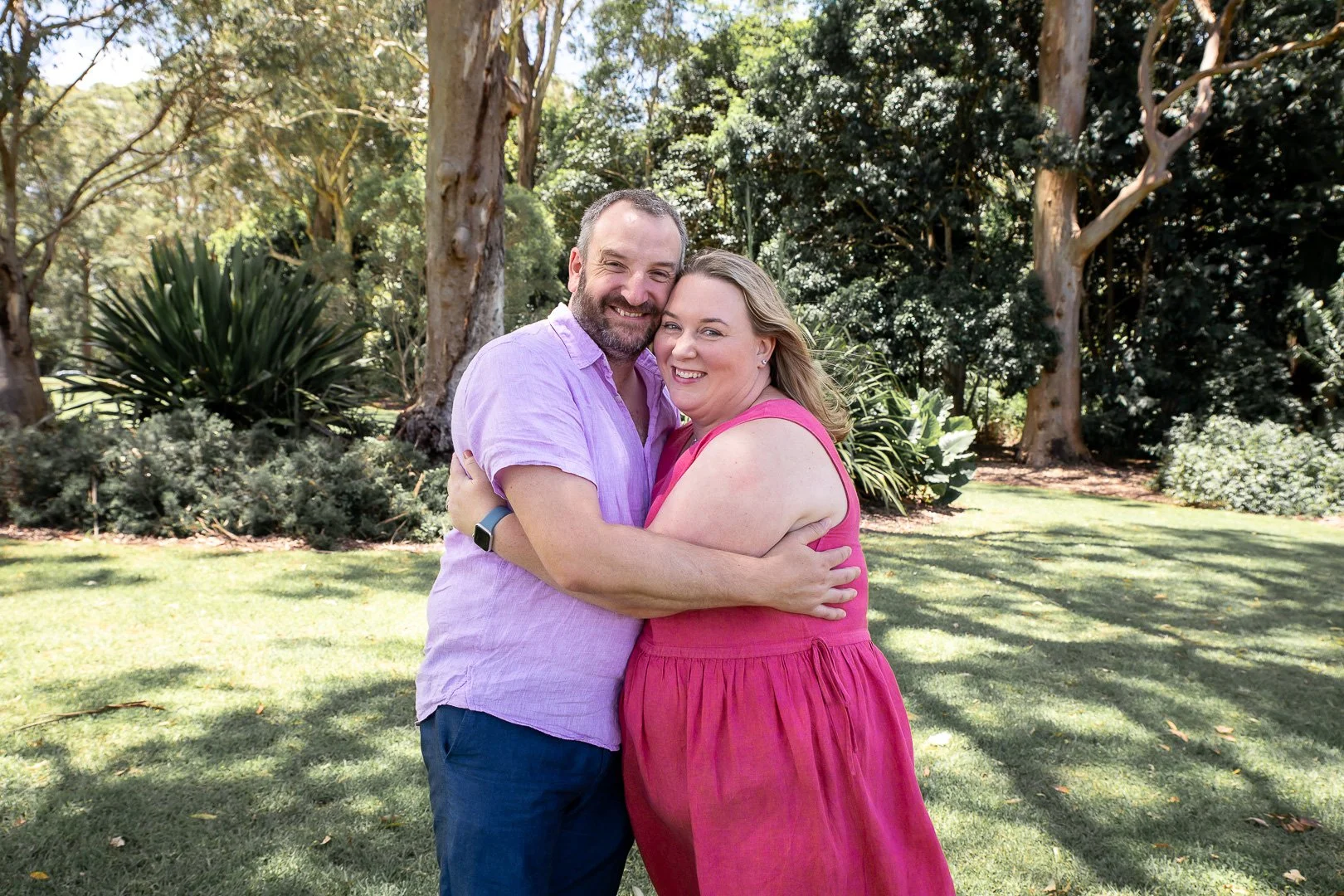 A couple embracing outdoors, smiling at the camera. The man wears a light purple shirt and the woman wears a pink sleeveless dress. They stand on grass with trees and bushes in the background.