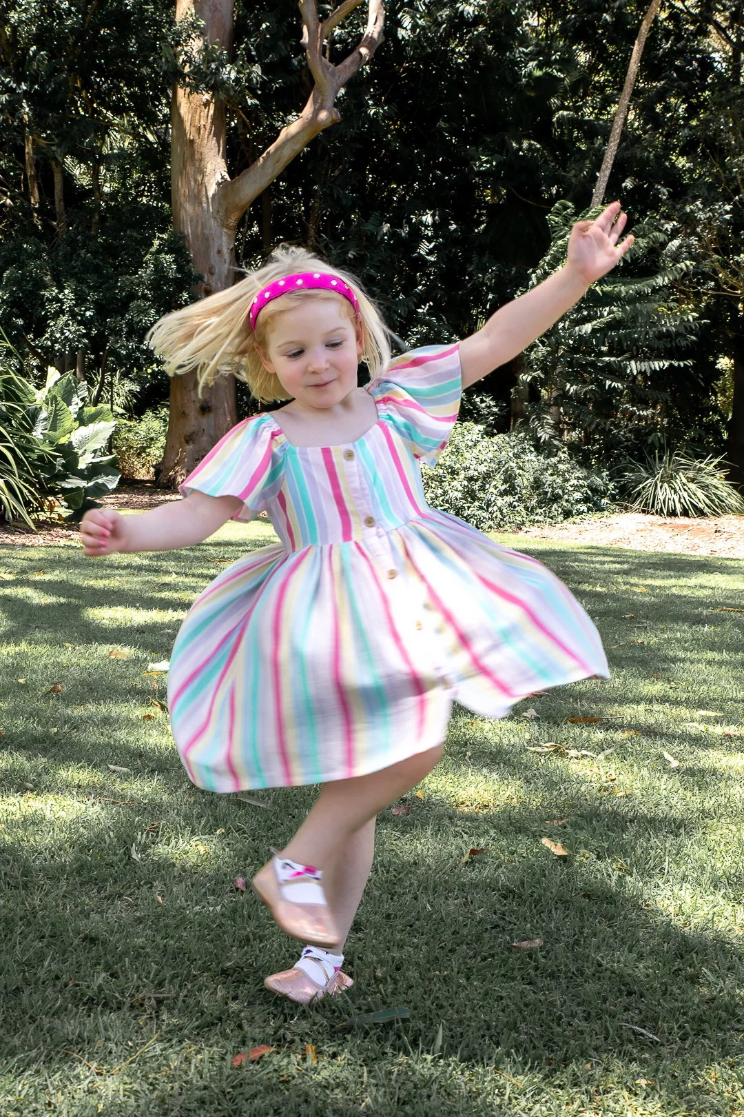 A young girl in a colorful striped dress twirling and jumping on a grassy lawn in a park with trees and bushes in the background.