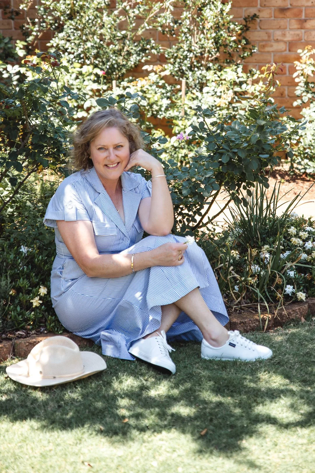 A smiling woman sitting on the grass in a garden with green bushes and a brick wall in the background, wearing a blue and white striped dress, white sneakers, and a bracelet, with a beige wide-brimmed hat on the ground beside her.
