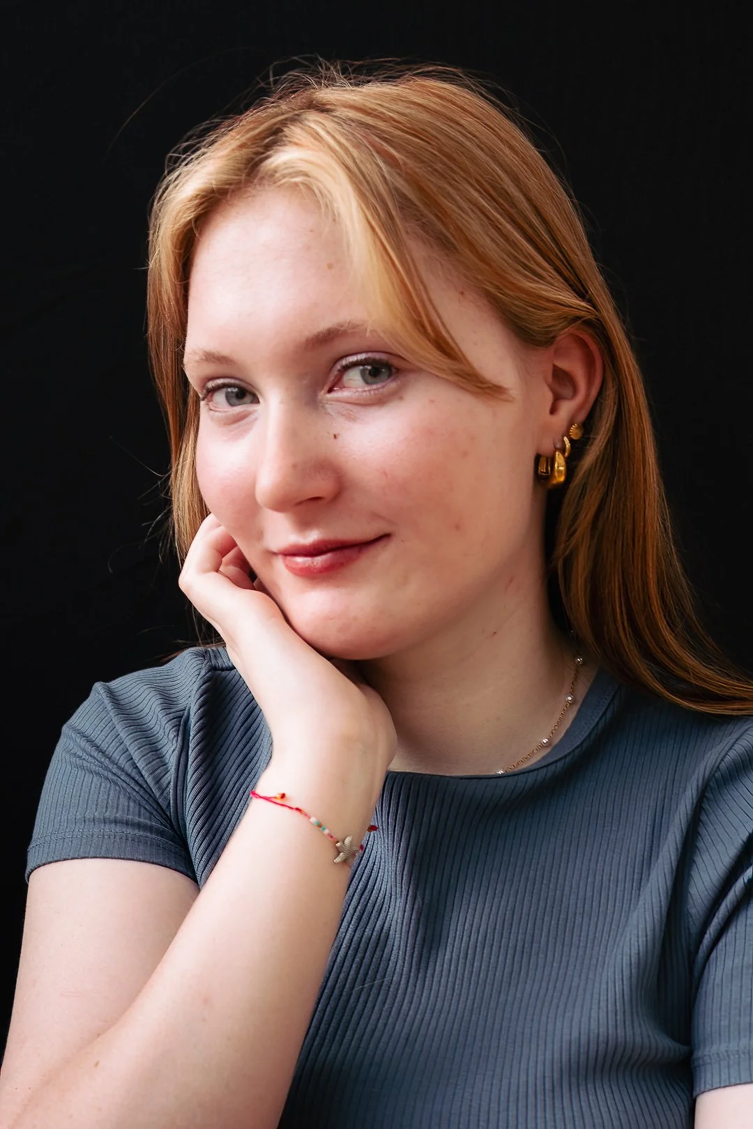 Close-up portrait of a young woman with ginger hair, blue eyes, and light freckles, wearing a gray top, jewelry, and a delicate smile, posed against a black background.