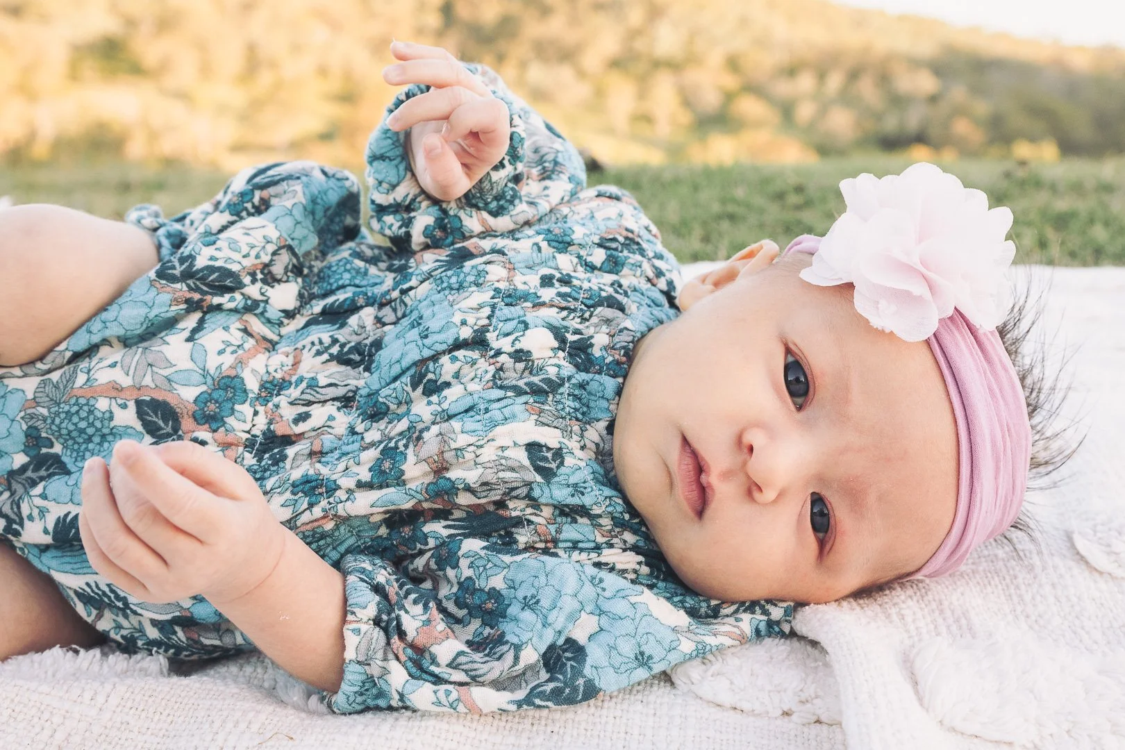 A baby girl lying on her side outdoors on a blanket, wearing a floral dress, pink headband with a large white flower, and a pink headband, looking at the camera with a peaceful expression.