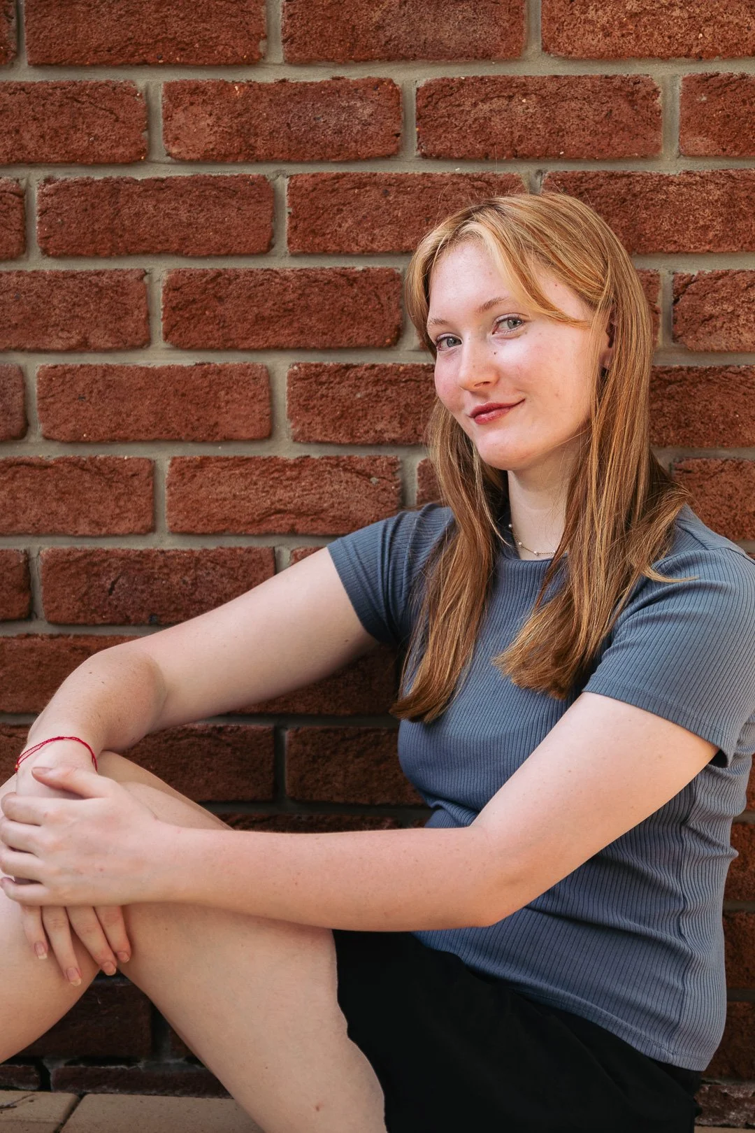 A young woman with long reddish hair sitting against a red brick wall, wearing a gray t-shirt and black shorts, looking at the camera with a slight smile.