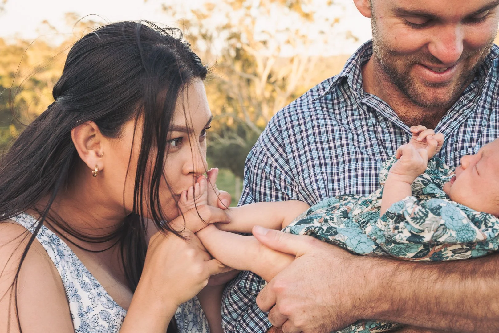 A woman and a man holding a newborn baby outdoors, with the woman gently kissing the baby's hand.
