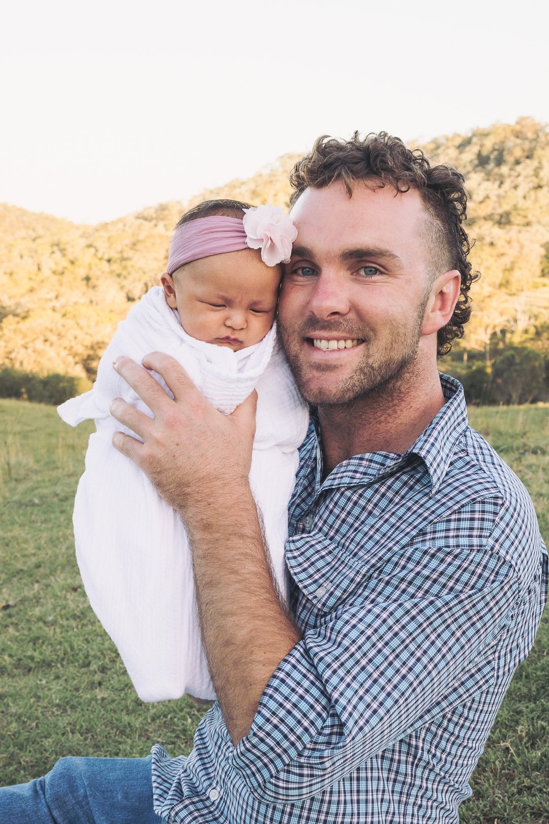 A man holding a sleeping baby girl outdoors, with a mountain landscape in the background.