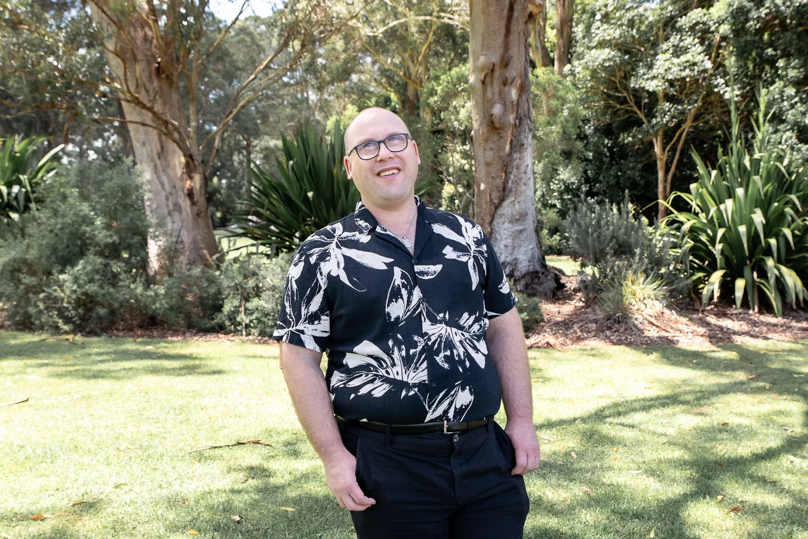 A man with glasses wearing a black and white floral shirt and black pants standing outdoors on grass with trees and plants in the background.