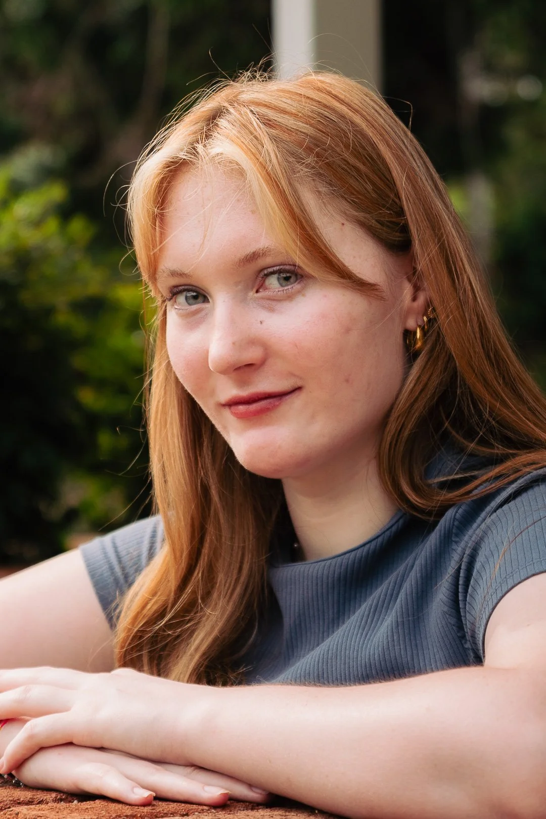 A young woman with red hair and blue eyes, smiling outdoors with green foliage in the background.