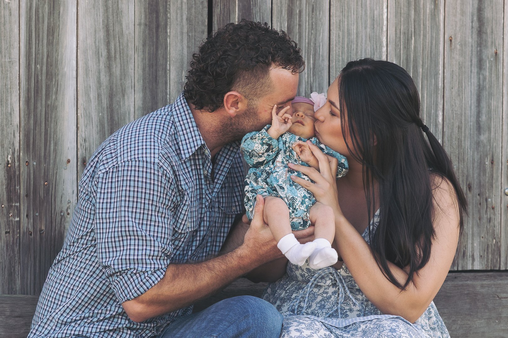 Family of three kissing and holding their baby girl in front of a wooden background.
