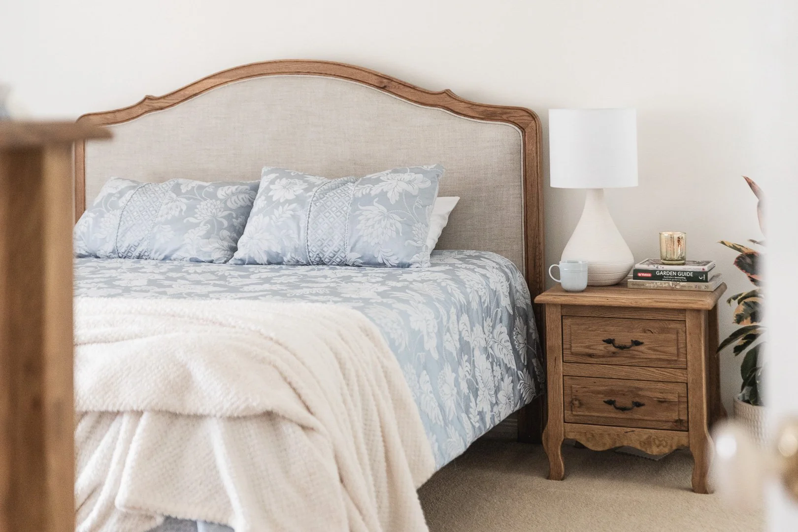 A bedroom with a bed that has a wooden headboard, light blue and white floral bedding, and three pillows. Next to the bed, there is a wooden nightstand with a white lamp, a white mug, a gold candle holder, and a couple of books including a garden guide. There are also some plants visible in the room.